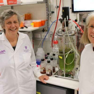 two women in white coats smile in a laboratory in front of a device containing green liquid